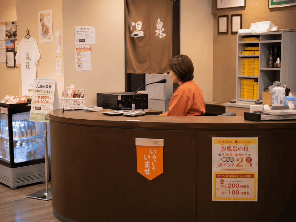 温泉施設のフロント（受付）で、スタッフの女性がマイクに向かって館内アナウンスをしゃっべてる様子。