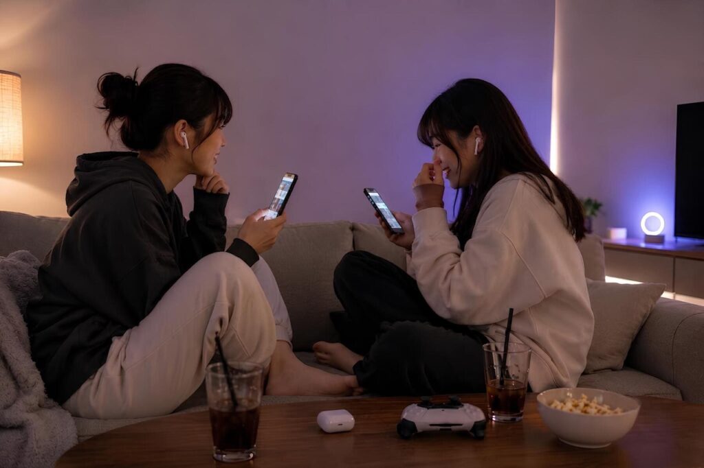 「夜、部屋のソファでスマートフォンを見ながら談笑する2人の若い女性。テーブルには飲み物やイヤホンケースが置かれ、リラックスした雰囲気が漂っている。」